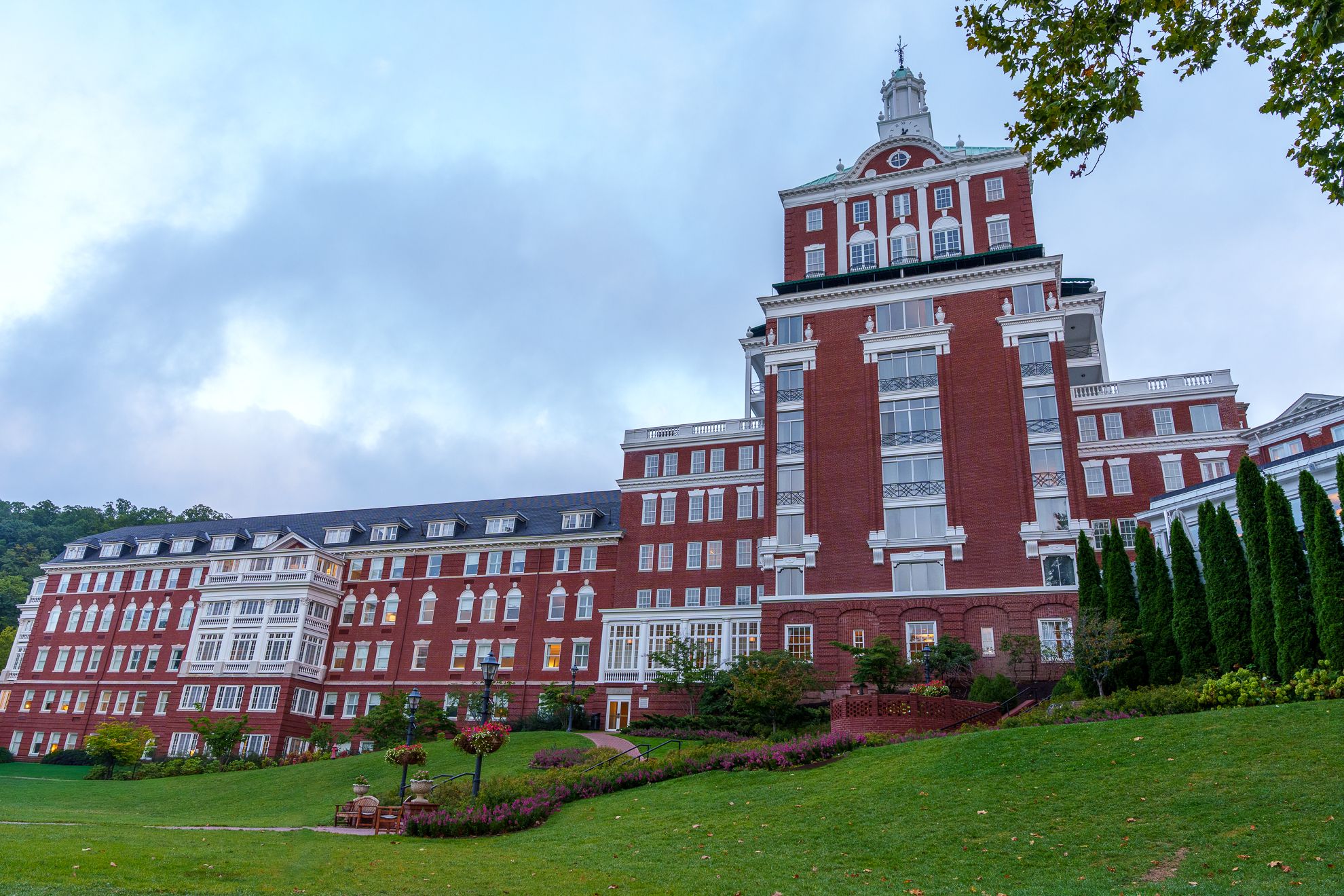 The rear of a grand red-brick resort hotel and its clock tower, seen across a lawn under a cloudy sky