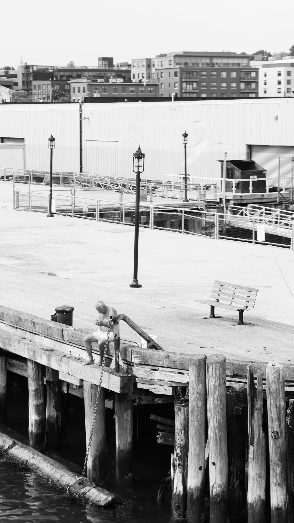 A figure seated at the edge of a weathered wharf, black and white